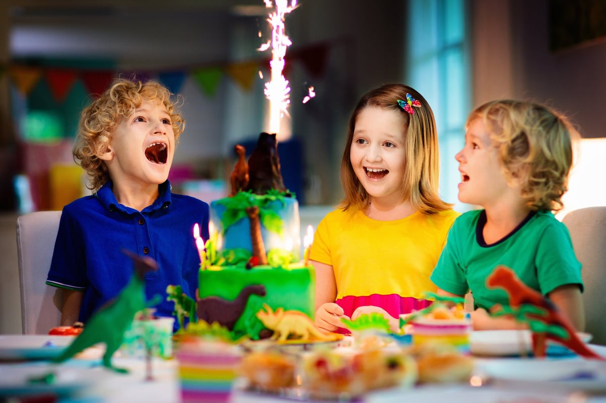 Kids birthday party with dinosaur theme cake, little girl blowing candles and opening gifts