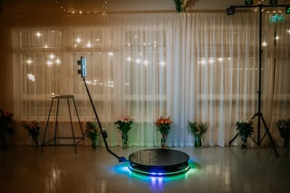 A 360-degree photo booth setup with LED lights and a rotating platform, surrounded by floral arrangements, in front of sheer curtains and string lights.