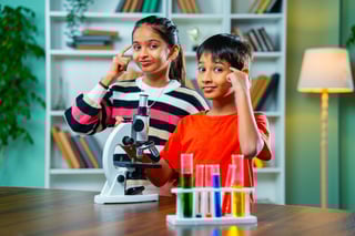 Indian Asian Curious Cute Little Girl and Boy students Sitting at Table in Modern Classroom, Playfully Studying Science Using Microscope
