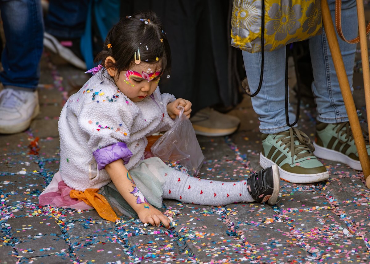 Little girl with face paint sits on the cobblestone street playing with colorful confetti at the Fasnacht carnival