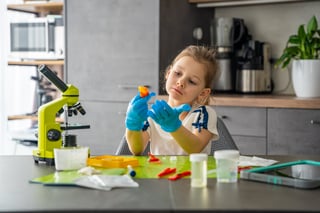 Girl wearing protective gloves examining grown crystal at table with microscope and laboratory tools