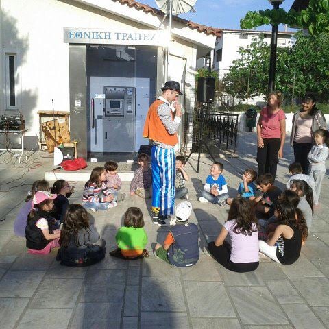 Street performer in colorful costume entertains a group of children sitting in a circle on a sunny plaza