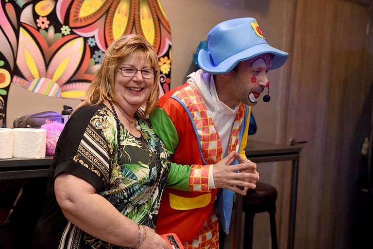 Woman in glasses and colorful shirt poses with man in blue baseball cap and clown costume in front of vibrant artwork