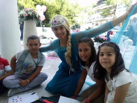 Adult dressed as Elsa posing with three smiling children at an indoor party or event with blue decorations and flowers.
