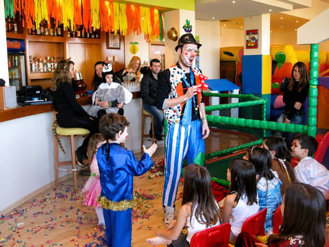 A colorful clown entertainer performs for a group of children in a bright indoor play facility with vibrant decorations and climbing structures