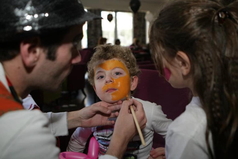 A toddler with yellow and orange face paint is held by two people during a makeup application session