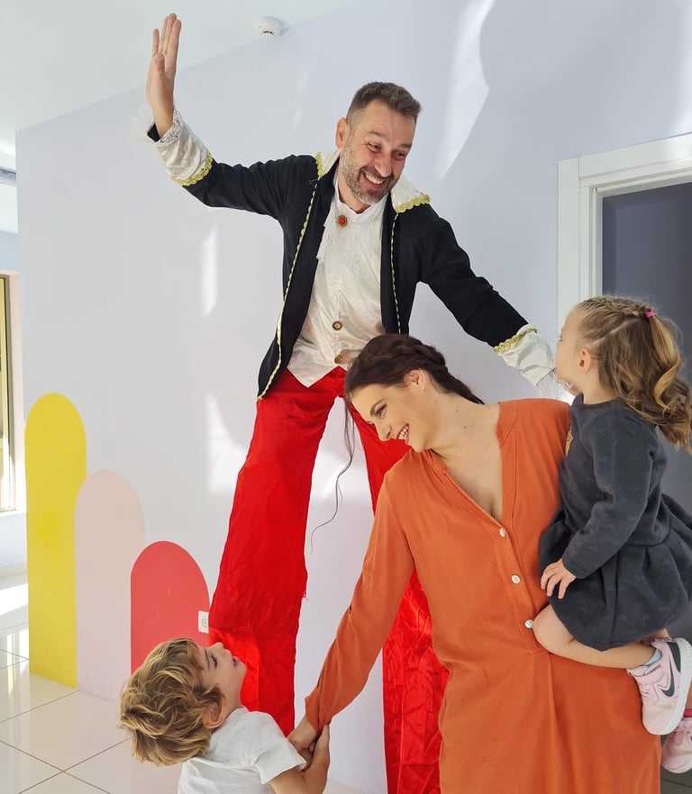 Man in formal jacket waving with arm raised, surrounded by woman in orange shirt and two children in white room with colorful shapes