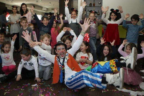 A group of children and an adult clown celebrate at an indoor party with colorful confetti on the ground.