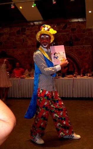 A performer dressed as a colorful clown in oversized clothing holding a childrens book at an indoor event