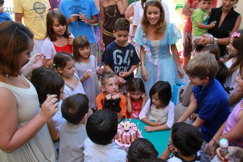 Group of children gathered around a birthday cake with an adult woman in the center at what appears to be an indoor childrens party or celebration event.