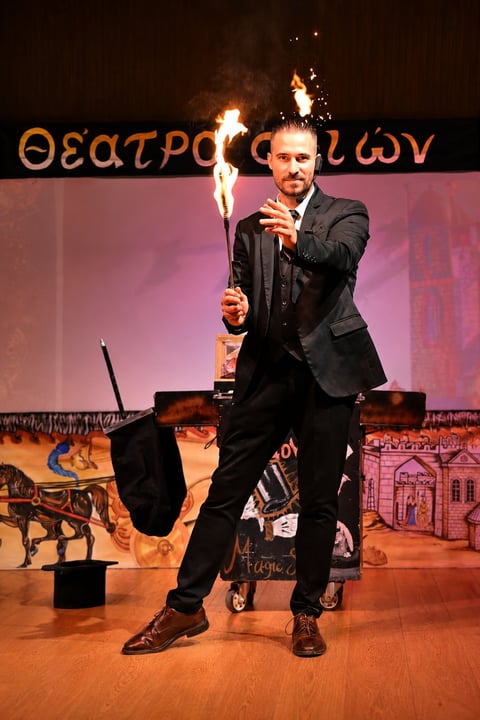 Man in black suit holding flaming torch on theatrical stage with Greek text backdrop