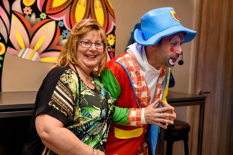 Woman in glasses and colorful shirt smiling next to clown performer in blue cap and checkered outfit at indoor event