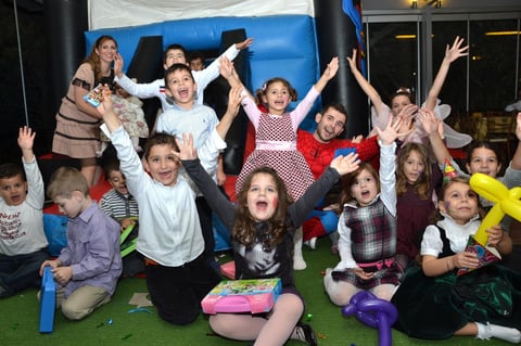 Group of children celebrating and posing with raised hands in front of a blue inflatable structure at an indoor party or event
